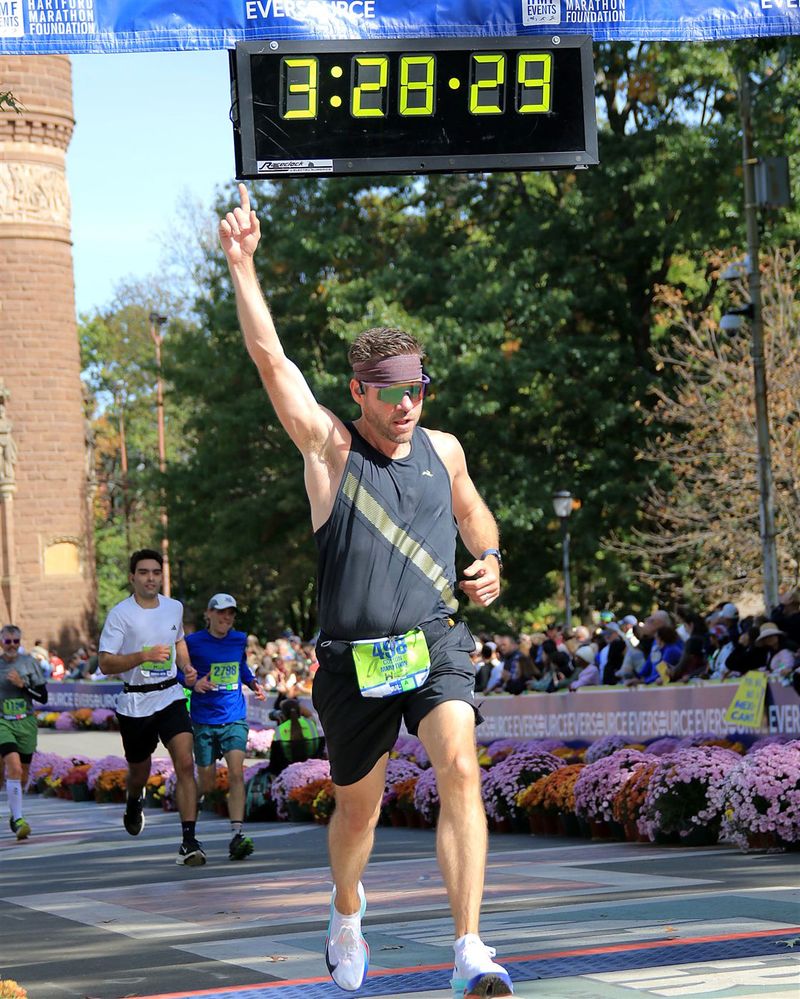 Colton crossing the Hartford Marathon finish line, arm raised, under the 3:28 race clock